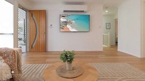 Een woonkamer met een grote flatscreen televisie aan de muur, een houten deur, een plant in een pot op een ronde mat en een kamerbreed tapijt met dessin.