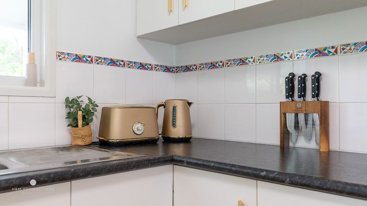 A kitchen with white cabinets, a black countertop, a gold-colored coffee maker, a kettle, and a knife block.