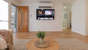A living room with a flat-screen TV mounted on the wall, a wooden door, and a potted plant on a round table. - Agnes Break | 3BR · Sauna & Walk to Beach (Agnes Water)