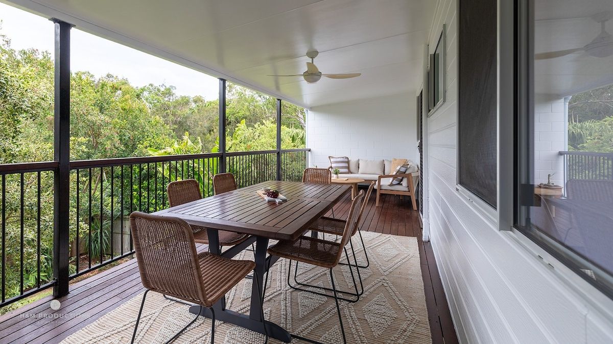 A covered balcony with a table and chairs, a sofa, and a view of greenery.
