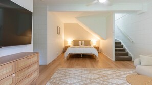 A bedroom with a bed, bedside lamps, a TV, a wooden dresser, and a staircase.