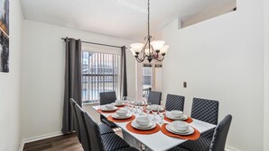 Un coin repas avec une longue table dressée pour six personnes, des chaises noires à revêtement en tissu et un lustre.