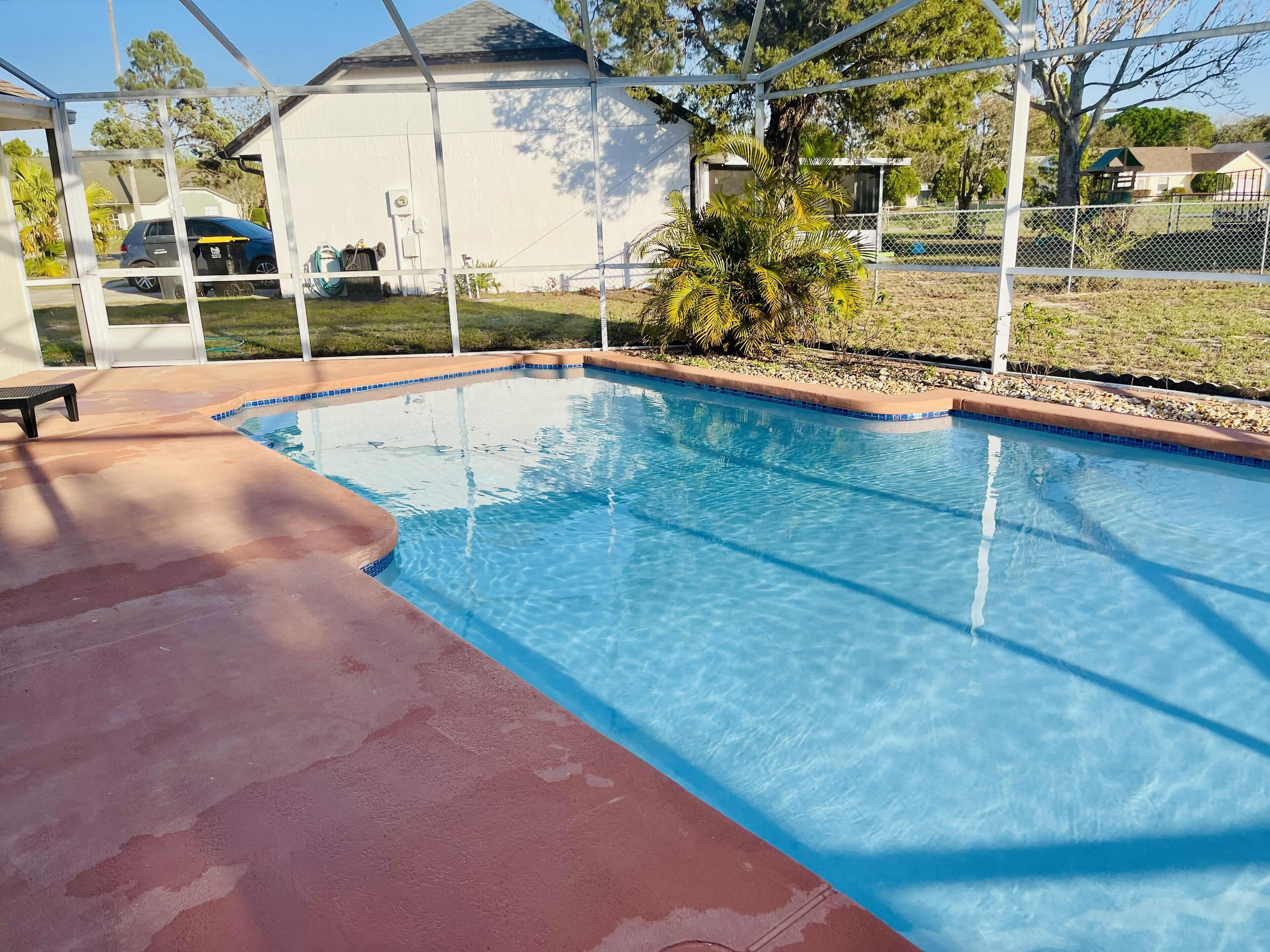 Una piscina con un deck rojo, rodeada por una cerca blanca y una pequeña palmera.