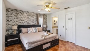 Une chambre avec un mur décoré d’un motif en briques, un lit avec une tête de lit, un ventilateur de plafond et une porte privative.