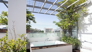 Une salle de bain extérieure moderne dotée d’une baignoire entourée de parois en verre, agrémentée de plantes en pot et surmontée d’une pergola équipée de panneaux solaires.