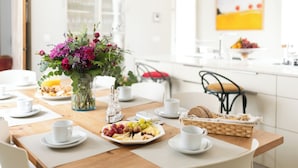 Une table de petit-déjeuner avec une table en bois, des chaises blanches, un vase de fleurs et une variété d’aliments pour le petit-déjeuner.