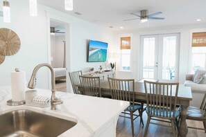 A modern kitchen with a sink, dining table, and chairs. There is a ceiling fan and a large window with blinds. - Enclave at Lyndell Lane by Avantstay Private Pool (Panama City Beach)