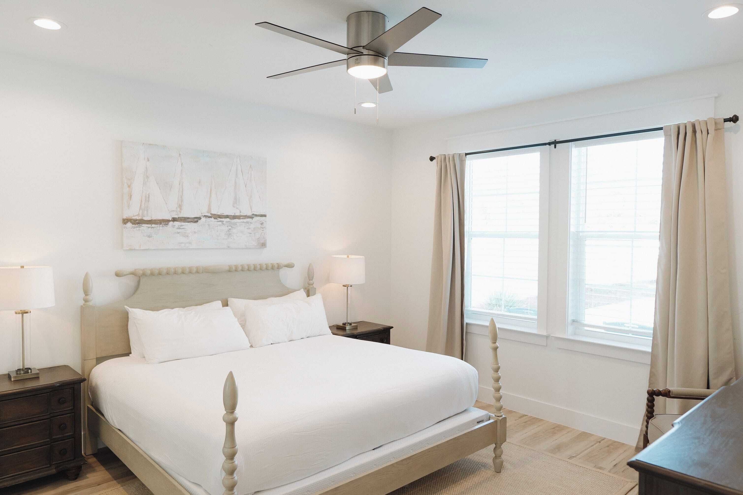 Une chambre à coucher bien rangée, avec un lit, des tables de chevet, un ventilateur de plafond et une grande fenêtre avec des rideaux.