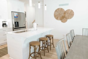 A modern kitchen with a large island, white cabinetry, stainless steel appliances, and a dining area with a wooden table and chairs. - Enclave at Lyndell Lane by Avantstay Private Pool (Panama City Beach)