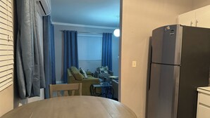 Kompakti oleskelutila, jossa on ruokapöytä, sohva, pieni pöytä tuolilla, jääkaappi ja ikkuna, jossa on verhot.