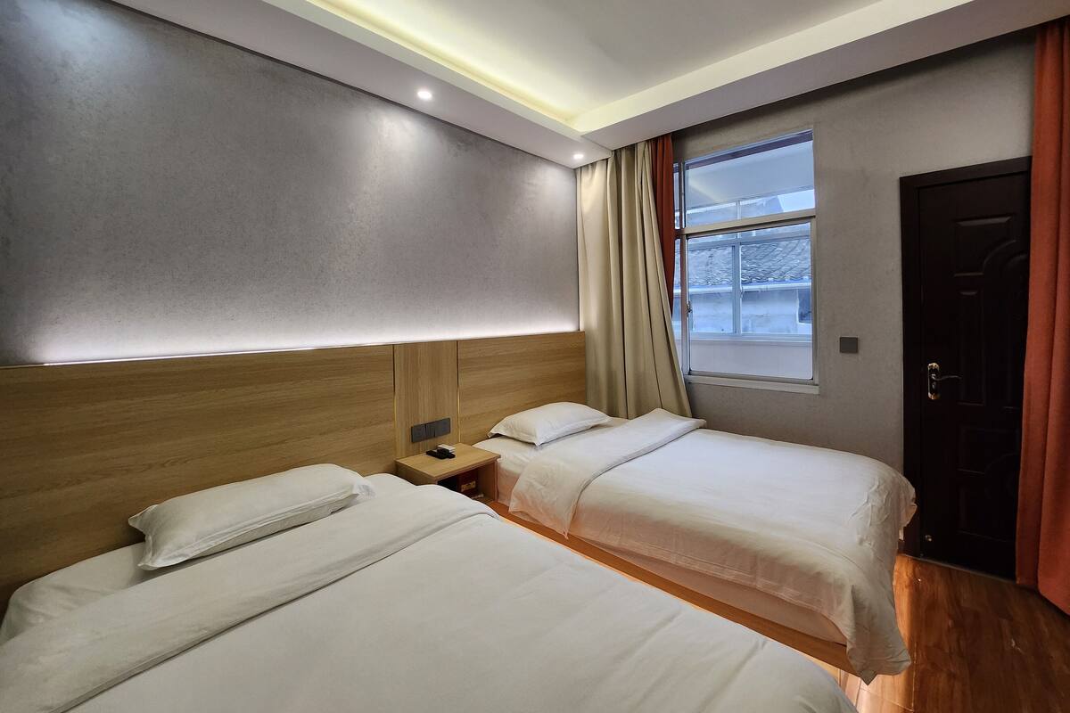 Habitación de hotel con dos camas, cabecera de madera, mesita de noche y ventana con cortinas.