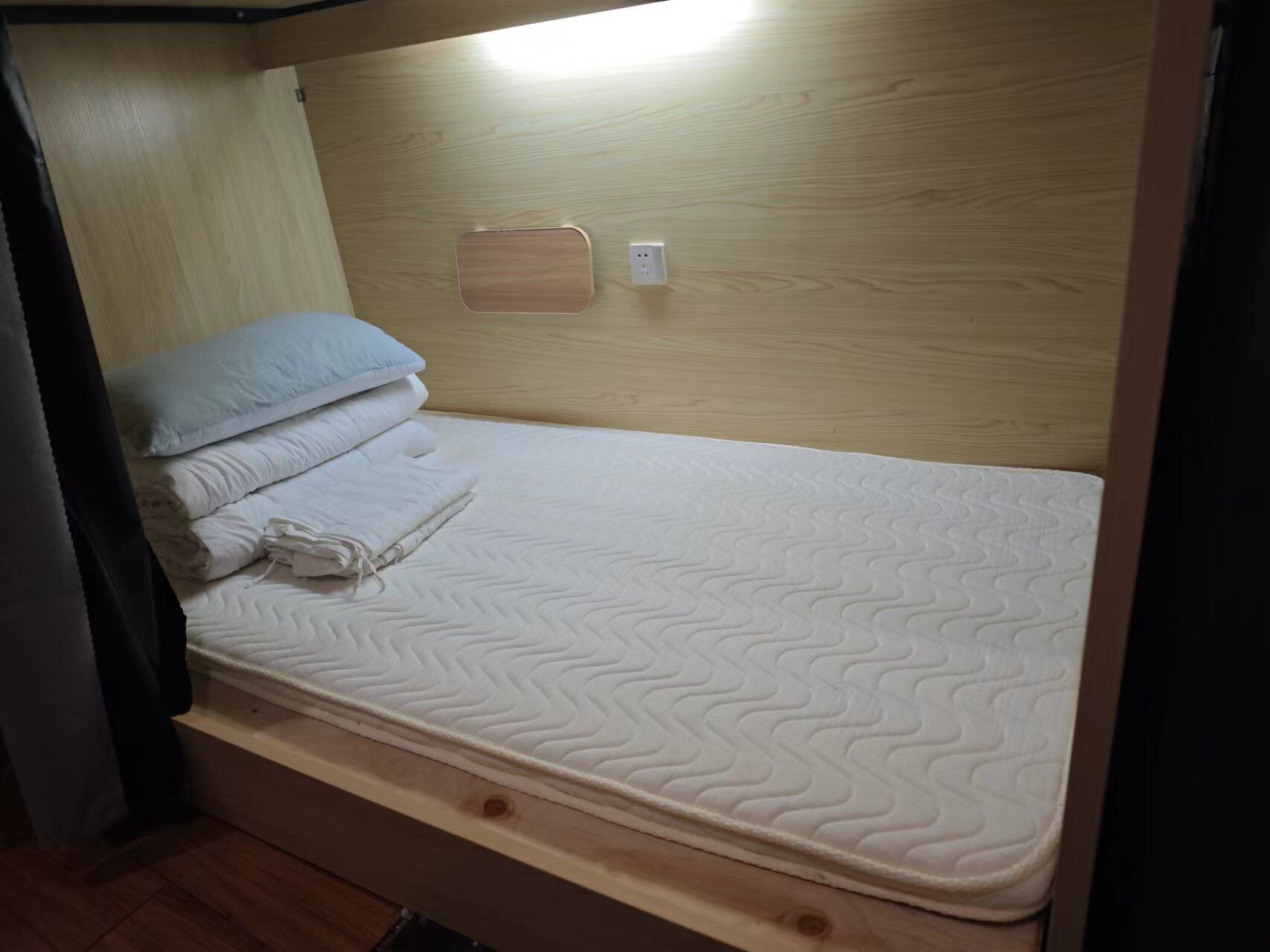A bunk bed with a mattress and pillows, a wooden headboard, and a power outlet on the wall.