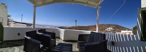 Ein Außenbereich mit Sitzgelegenheiten, Stühlen und einer Pergola mit Blick auf eine Hügellandschaft und das Meer.