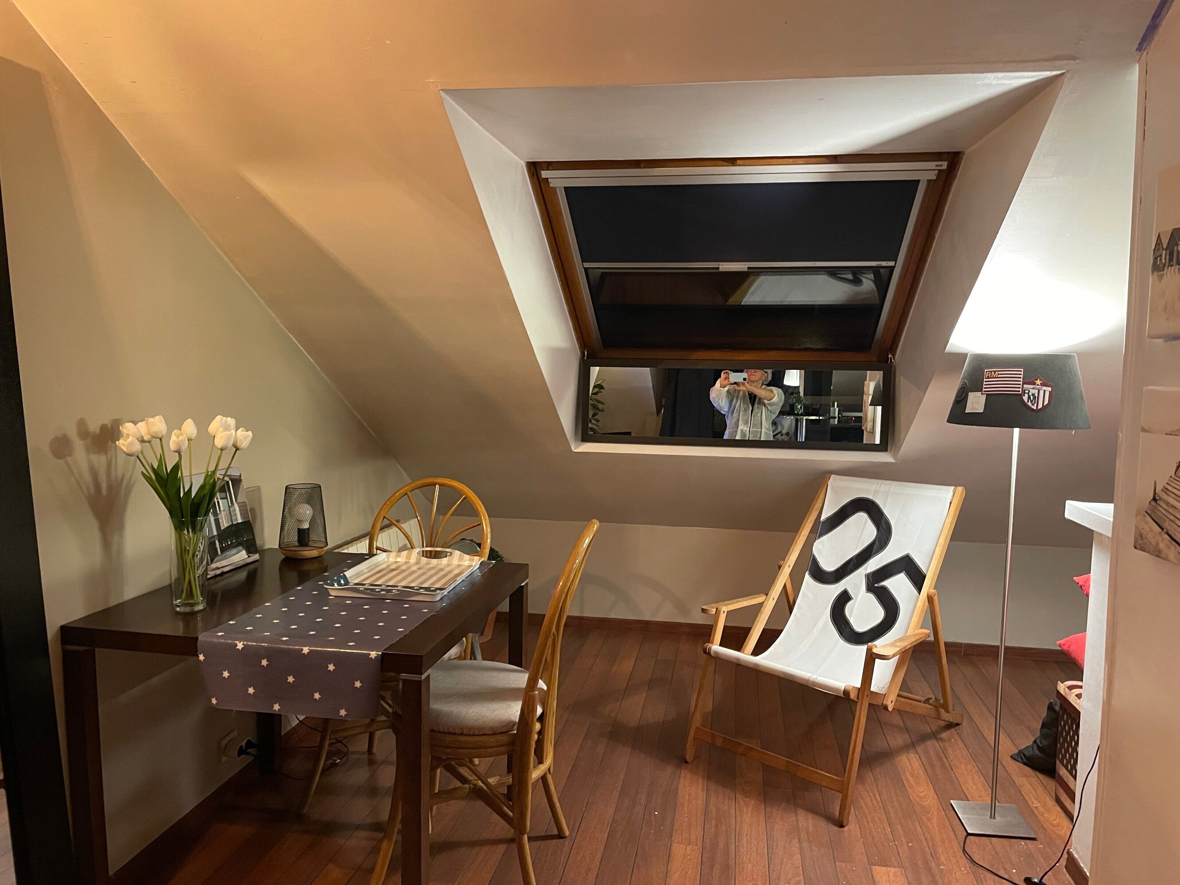 Ein kleiner Raum mit schrägverdeckter Decke, einem Esstisch mit Stühlen, einem Fenster und einer Lampe.