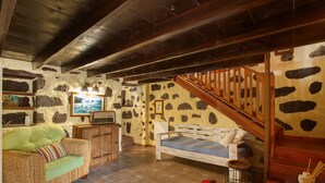 A rustic room with a stone wall, wooden ceiling beams, a wooden staircase, a bed, a sofa, and a television.