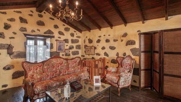 A room with a stone wall, wooden ceiling, a chandelier, a sofa, a chair, and a glass coffee table.