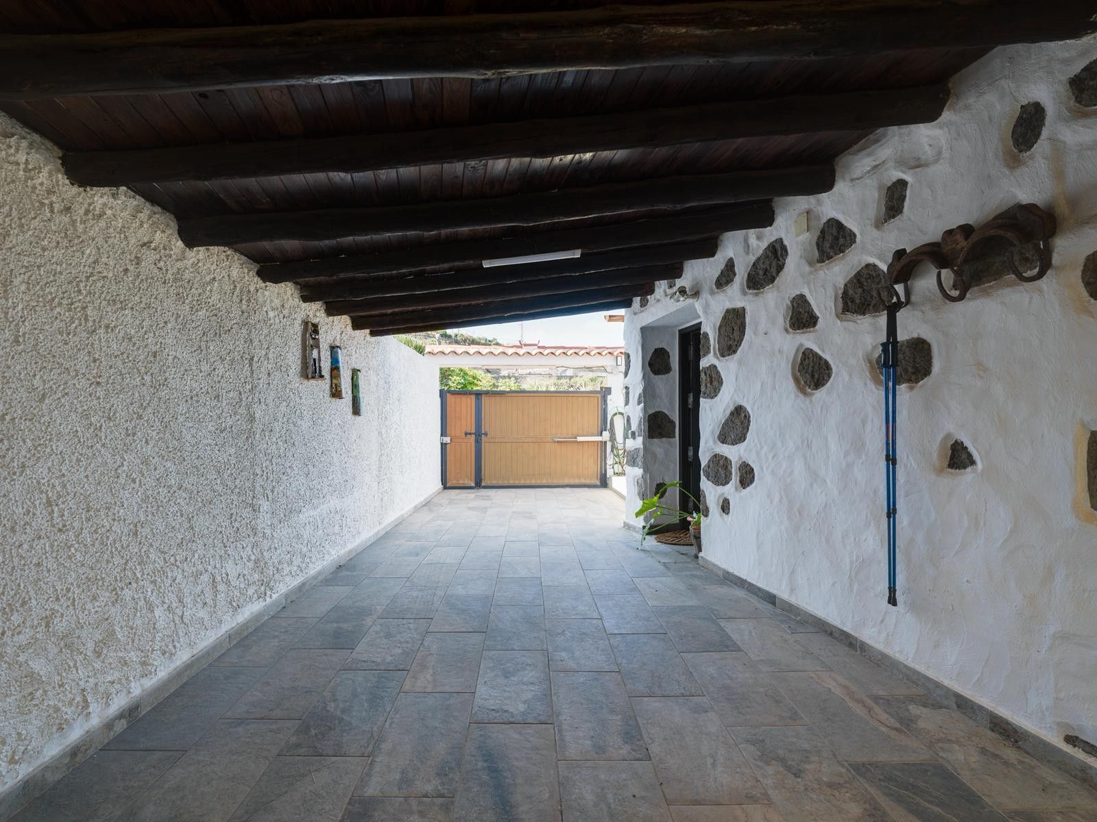 A corridor with a stone wall on one side and a wooden ceiling on the other, leading to a wooden gate.