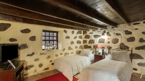 Une chambre avec des murs en pierre, des poutres en bois, deux lits, une télévision et un tapis rouge.
