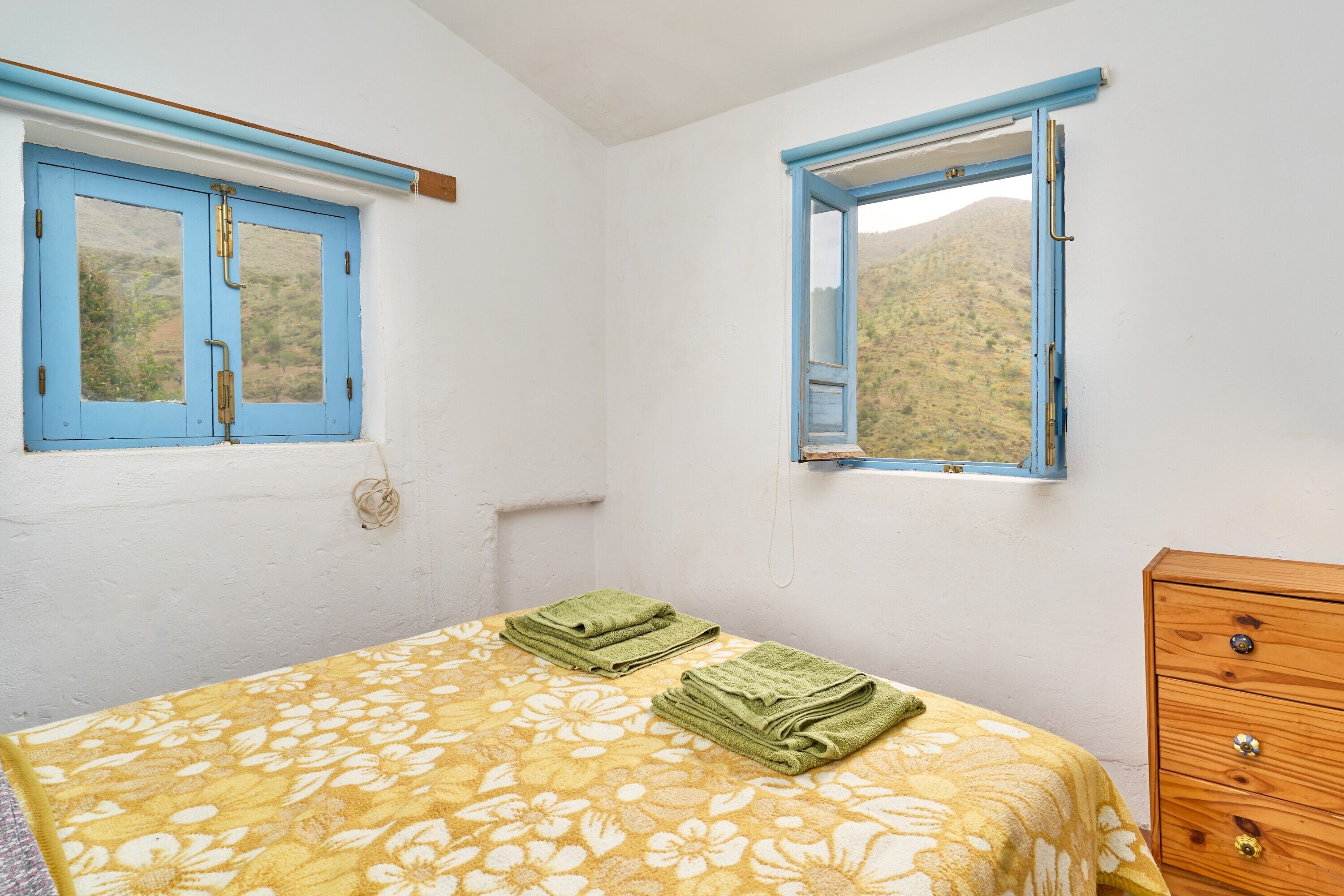 Une chambre à coucher avec un lit, deux fenêtres, une commode en bois et une vue sur un paysage vallonné.