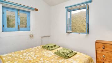 Une chambre à coucher avec un lit, deux fenêtres, une commode en bois et une vue sur un paysage vallonné.