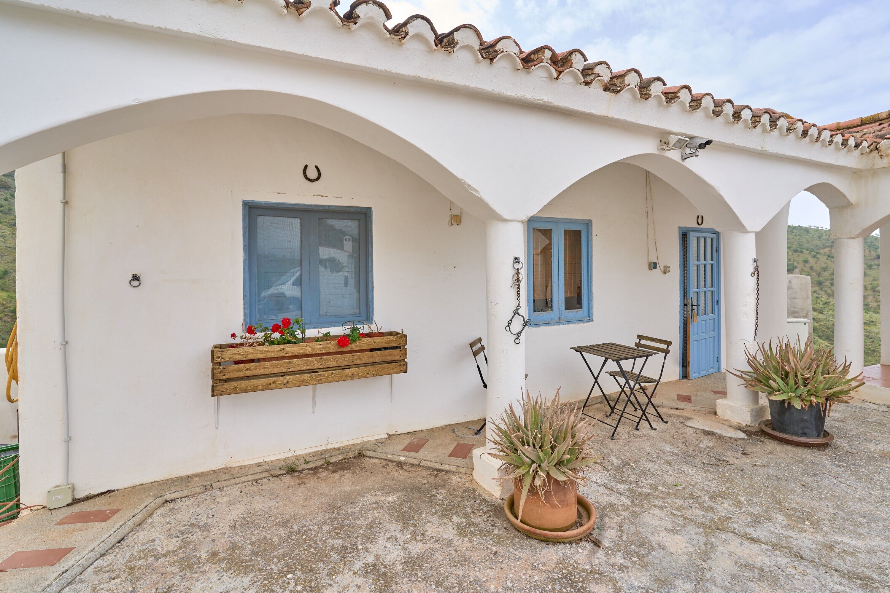 Un bâtiment blanc avec des fenêtres et des portes à encadrement bleu, un banc en bois orné de fleurs, une table et des plantes en pot.