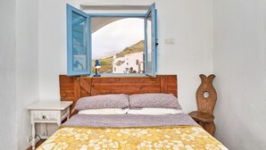 A bedroom with a wooden headboard, a bed with floral bedding, a nightstand, a blue lamp, and a view of a white building through an open window. - Country House 'The Tack Room' with Mountain View, Shared Pool and Wi-Fi (Rubite)