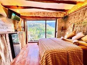 A bedroom with a bed, a TV, a small table, and a view of a forested area. - Apartment 'Duende' with Mountain View, Balcony and Air Conditioning (Olba)
