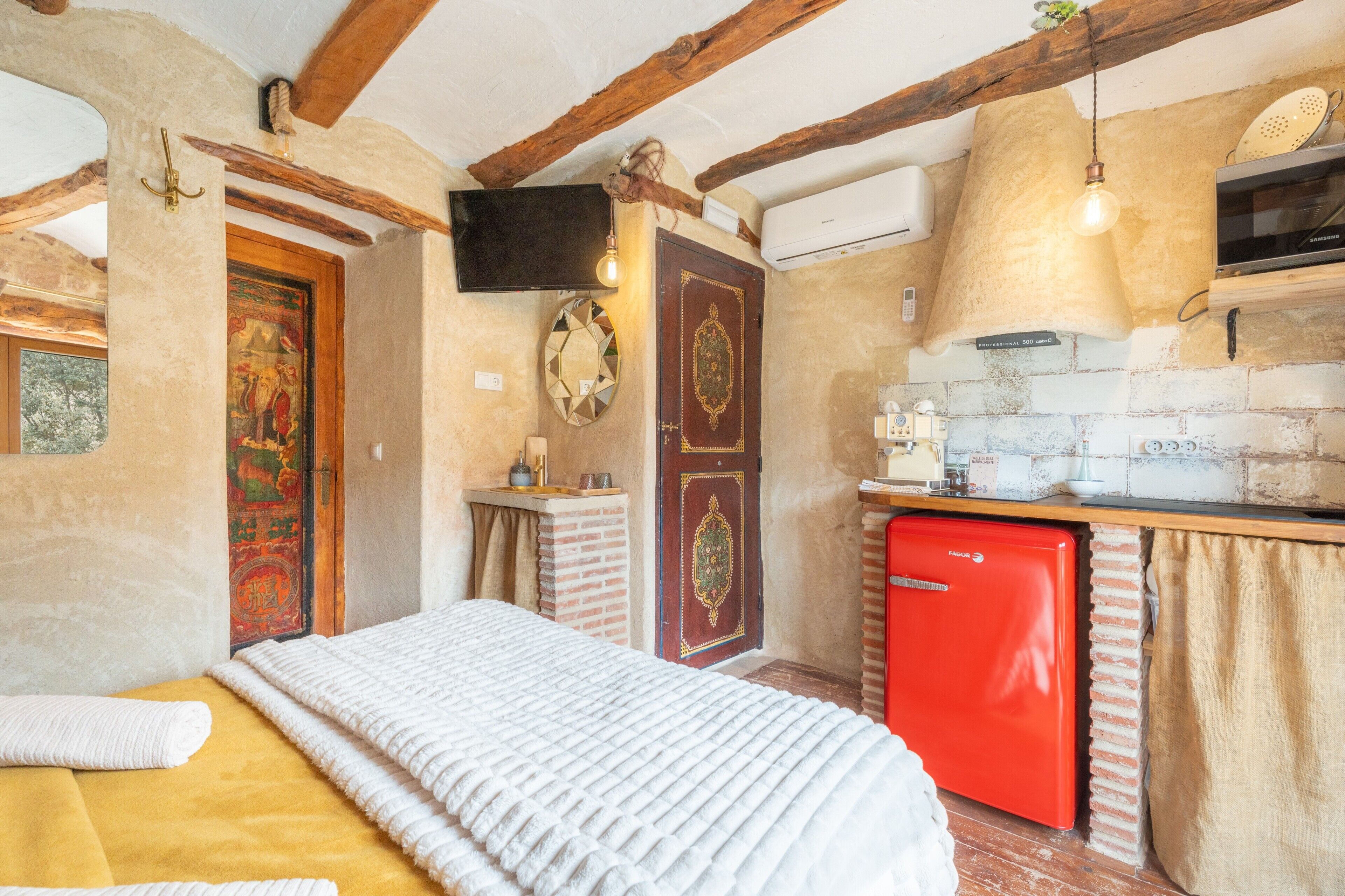 A rustic room with a bed, a red refrigerator, a television, and a kitchen area with a brick wall and wooden beams.