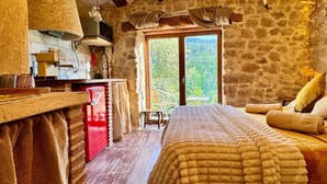 Ein rustikaler Raum mit freiliegenden Holzbalken, einer Steinwand, einem Bett mit strukturierter Decke, einem roten Schrank und einem Fenster mit Blick auf Grünflächen.