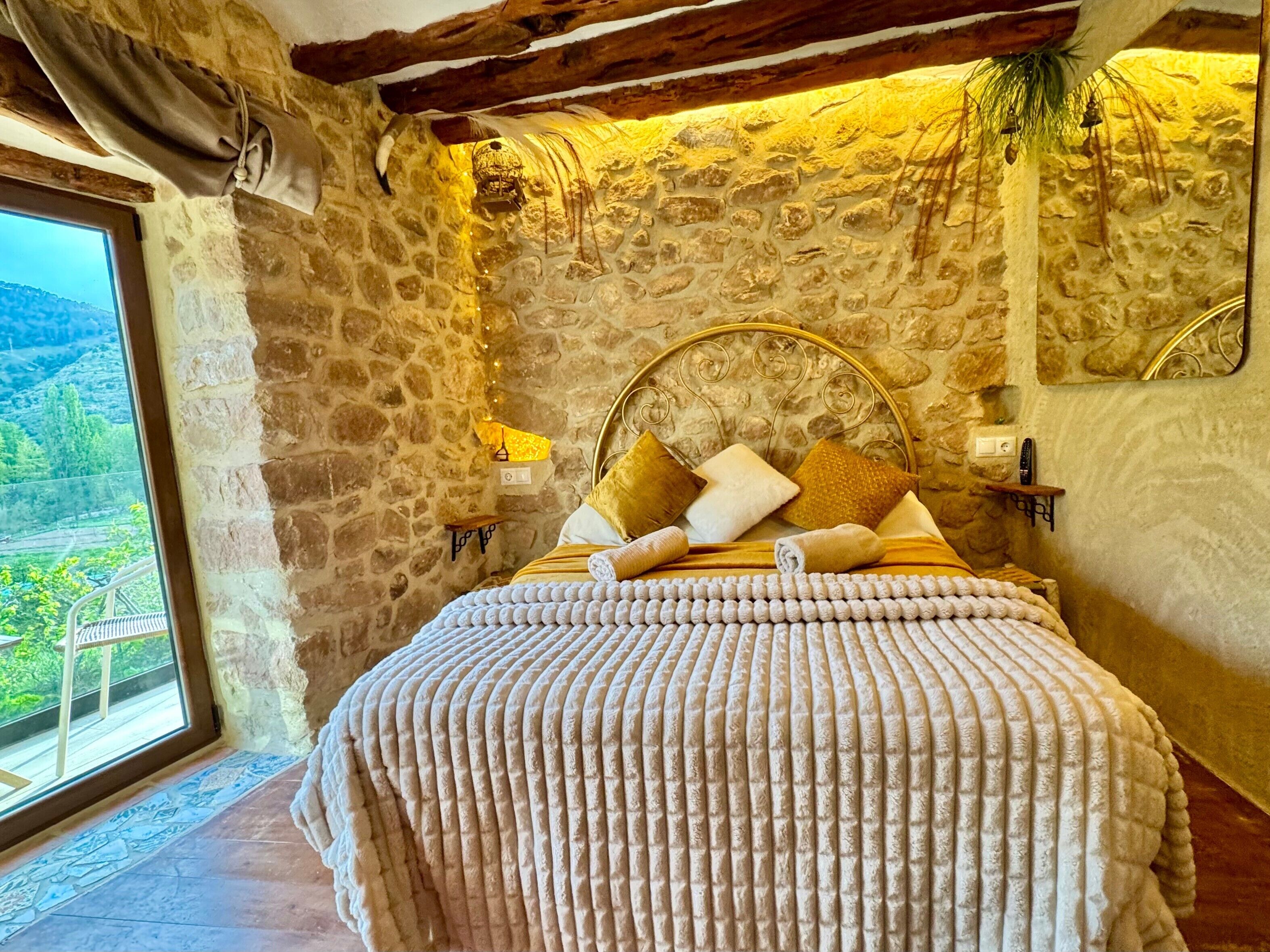 A stone-walled bedroom with a bed, pillows, a headboard, a nightstand, a lamp, and a view of greenery outside.