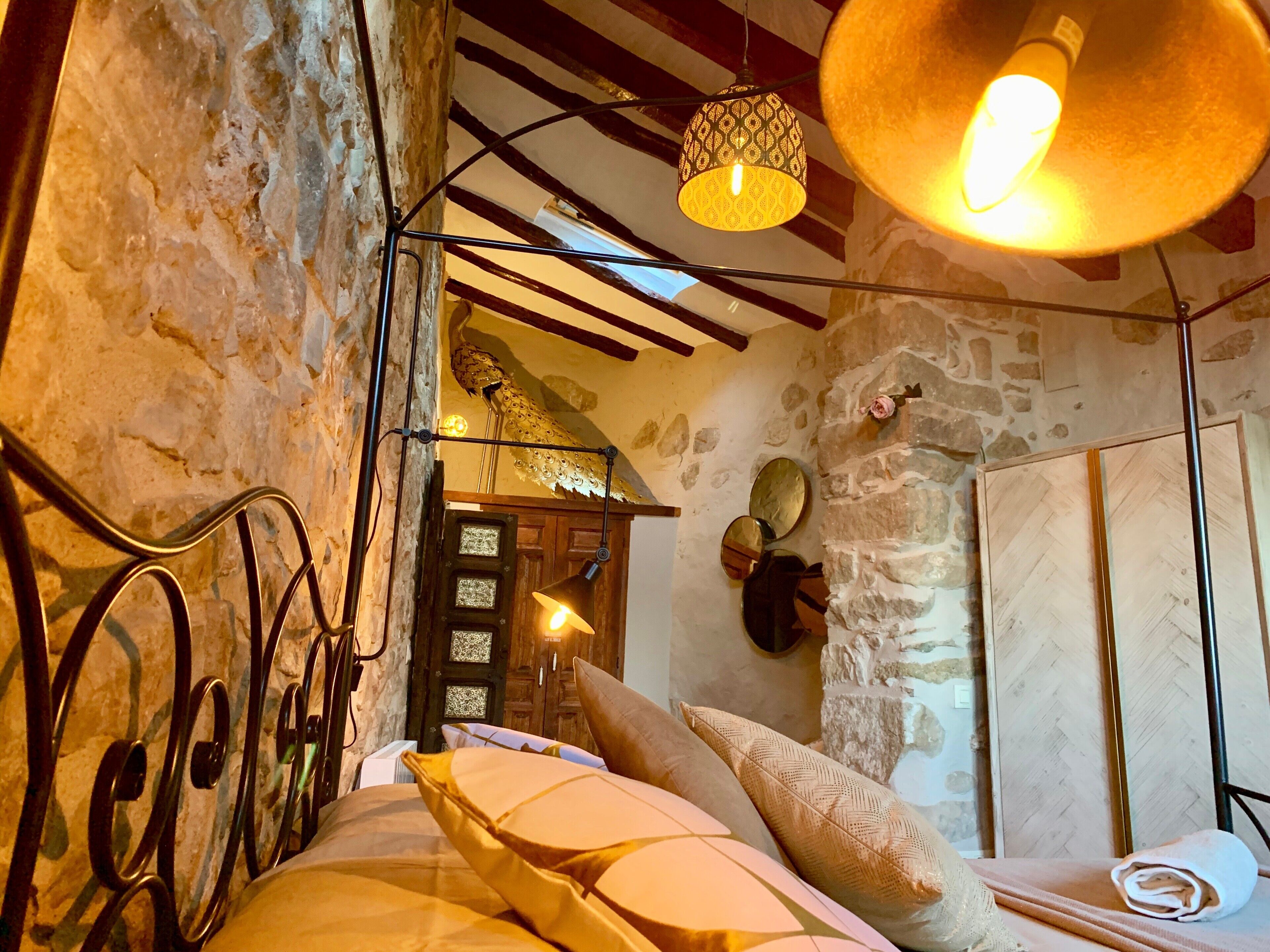 A rustic room with a stone wall, a wooden door, a bed with pillows, a hanging light fixture, and a mirror on the wall.