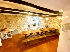 Ein Zimmer mit Steinwänden, einer Holzbänkl und einem Tisch, einem kleinen weißen Tisch und einem Korb auf der linken Seite.