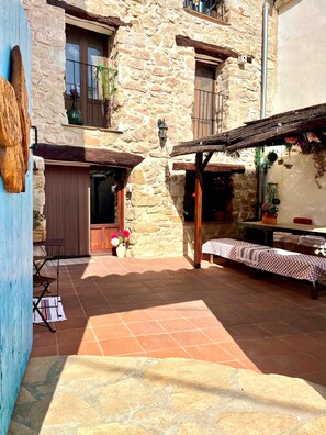 Ein Steinhaus mit einer Terrasse, einer hölzernen Bank und einem Tisch mit kariertem Tischtuch.