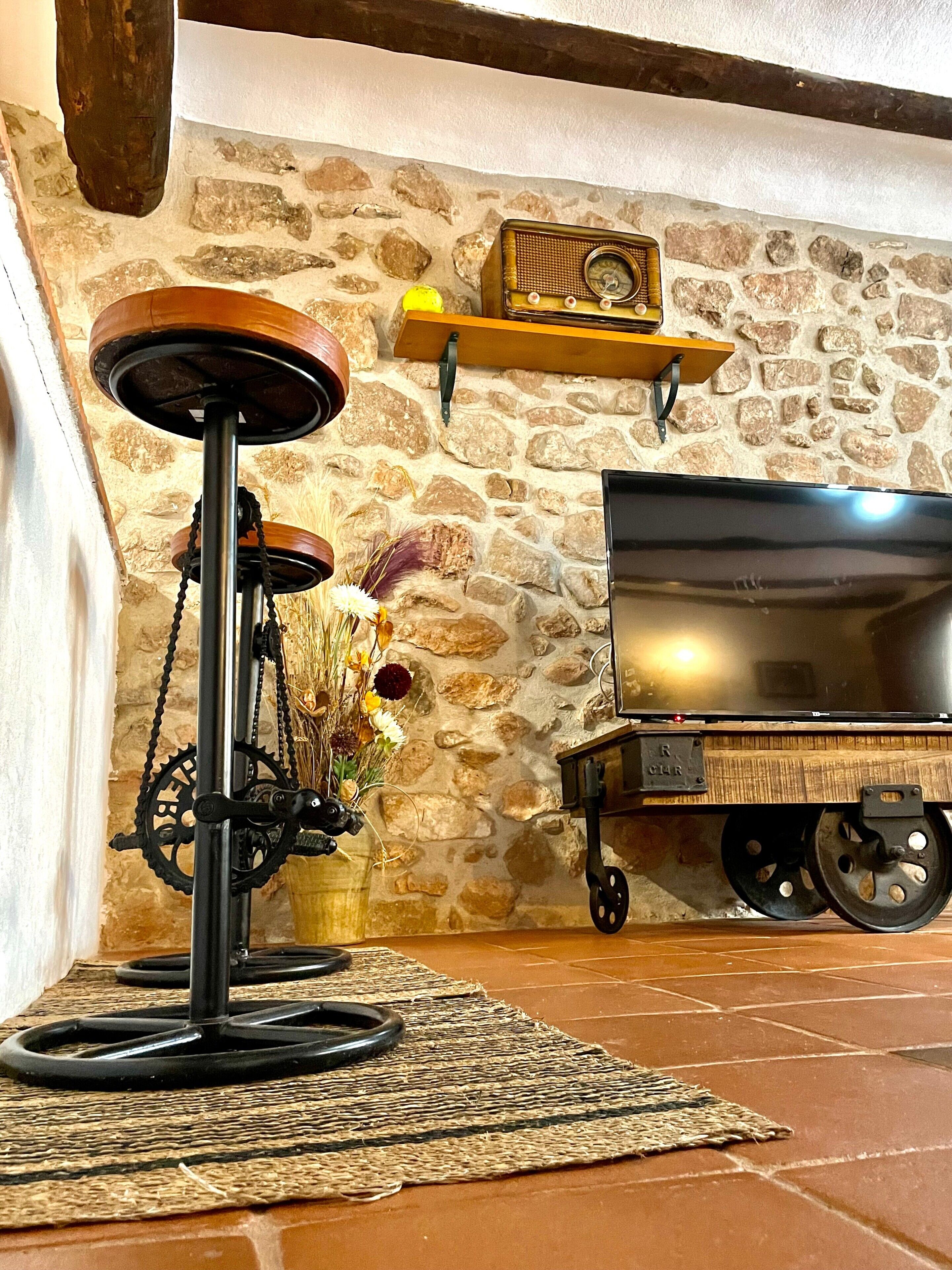 Un intérieur rustique avec un mur en pierre, une étagère en bois sur laquelle est posée une horloge, un tabouret de bar ancien et un chariot à roulettes.
