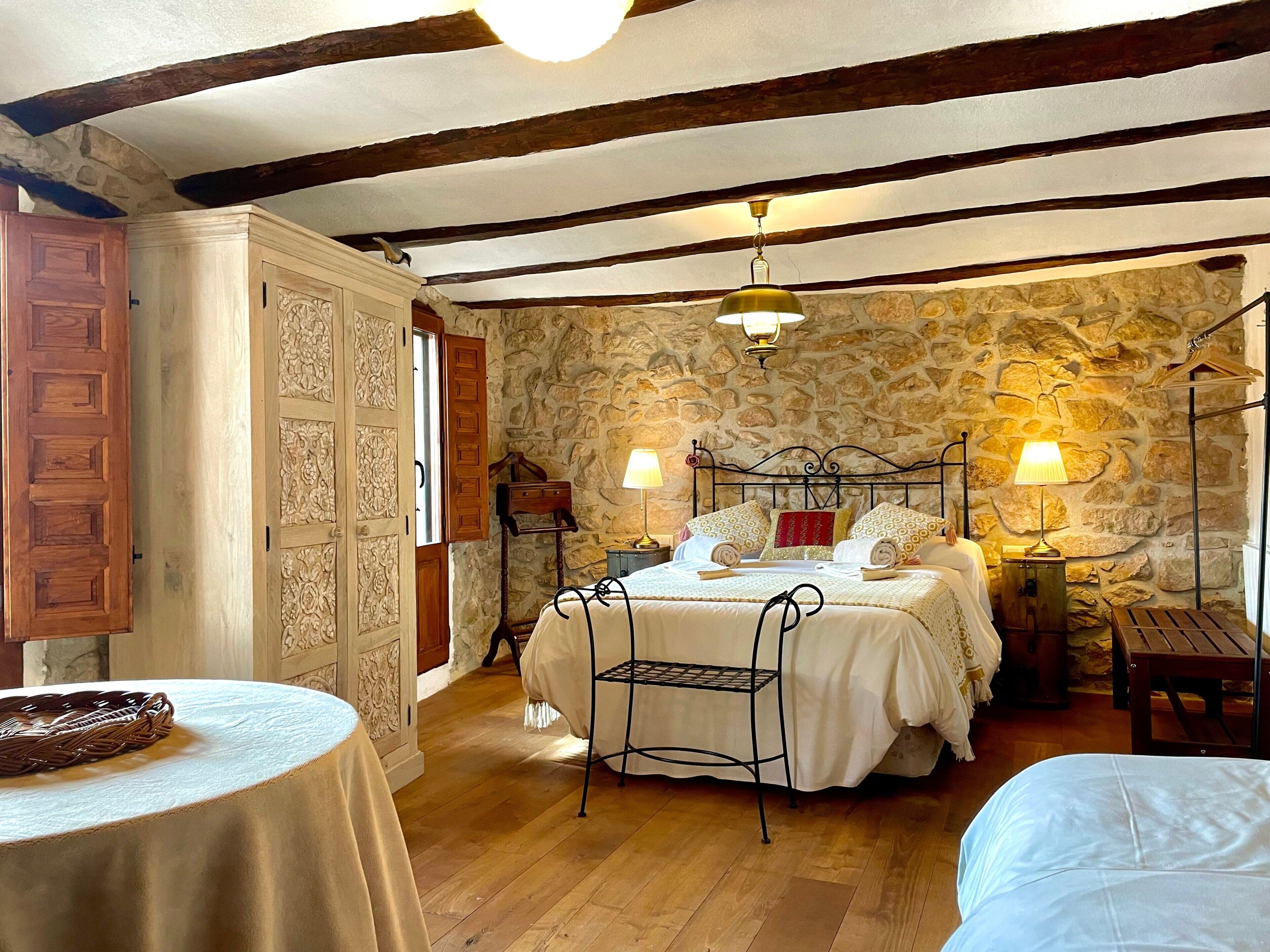 A bedroom with a stone wall, a bed with two pillows, a nightstand, a lamp, and a chair.