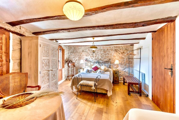 A bedroom with a stone wall, wooden beams, a bed with pillows and a blanket, a bench, a table with a basket, and a hanging lamp.