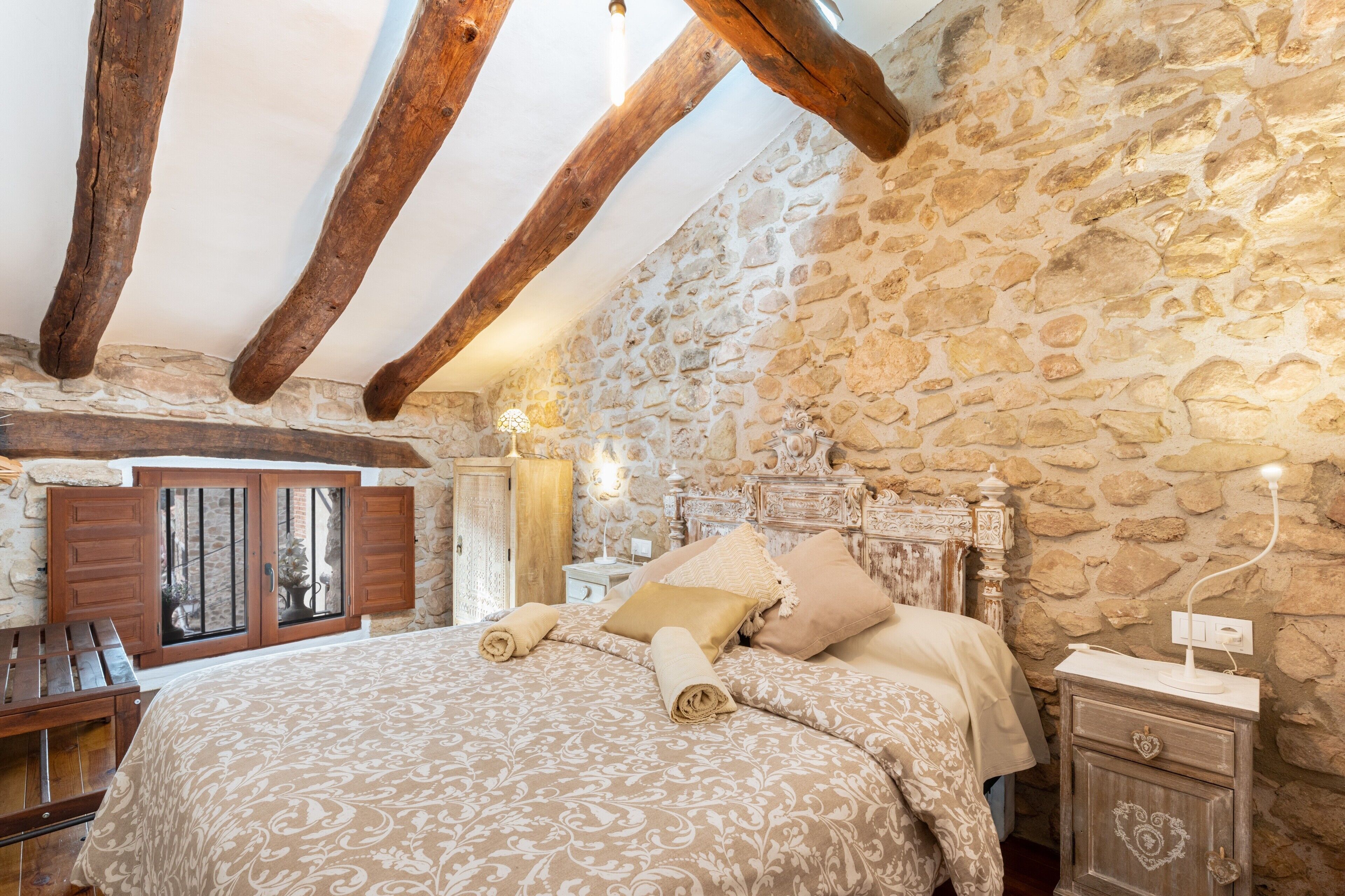 Un dormitorio con pared de piedra, vigas de madera, una cama con toallas enrolladas, una mesita de noche y una lámpara.