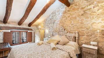 Un dormitorio con pared de piedra, vigas de madera, una cama con toallas enrolladas, una mesita de noche y una lámpara.