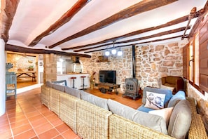 A rustic interior with exposed wooden beams, a stone wall, a stone fireplace, a wicker sofa, and a television.