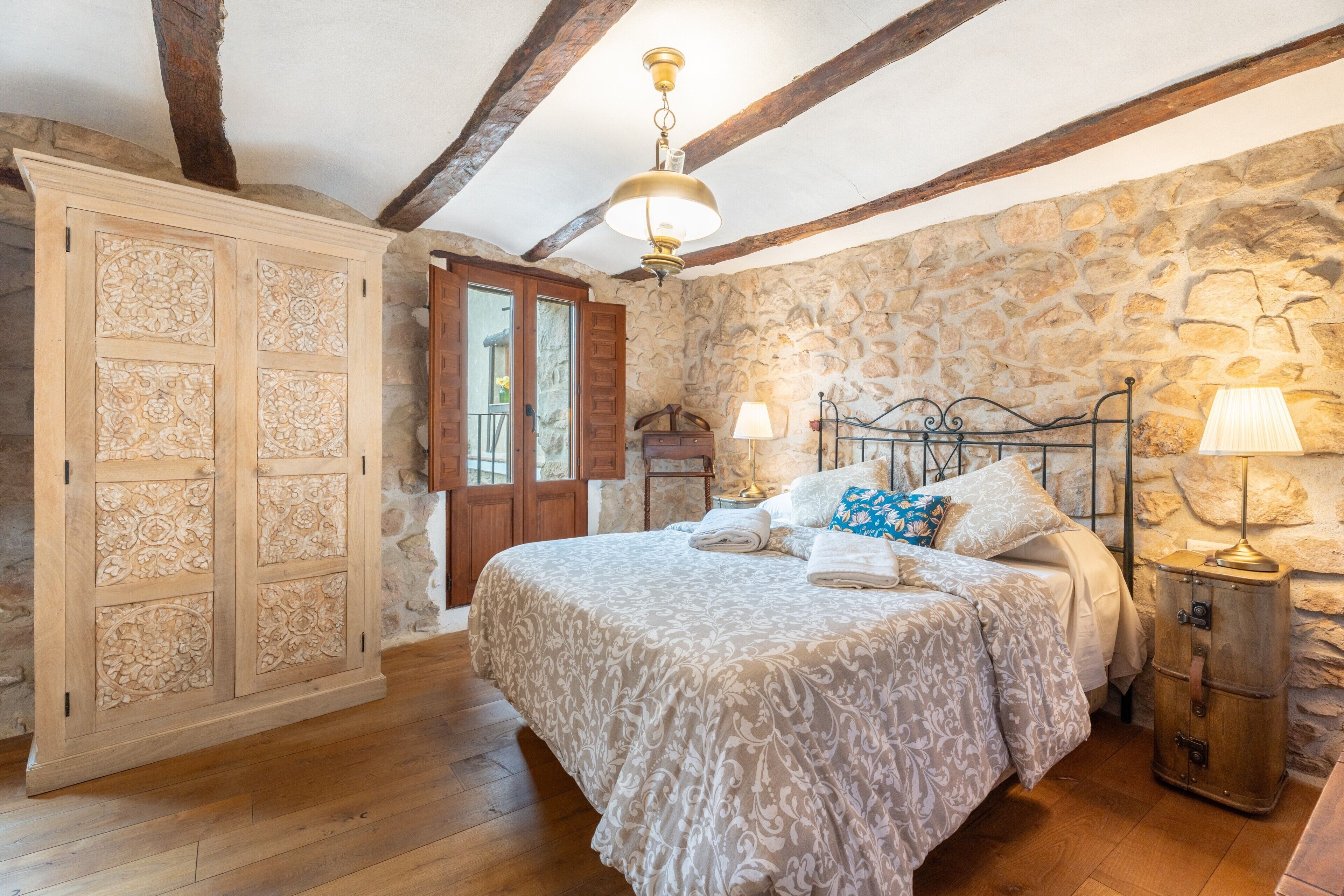 A bedroom with a stone wall, wooden ceiling beams, a bed with patterned bedding, a wardrobe, and a nightstand with a lamp.