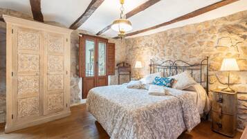 A bedroom with a stone wall, wooden ceiling beams, a bed with patterned bedding, a wardrobe, and a nightstand with a lamp.