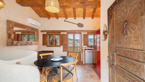 Um interior rústico com área de jantar, vigas de madeira no teto e cozinha visível através de uma porta aberta.