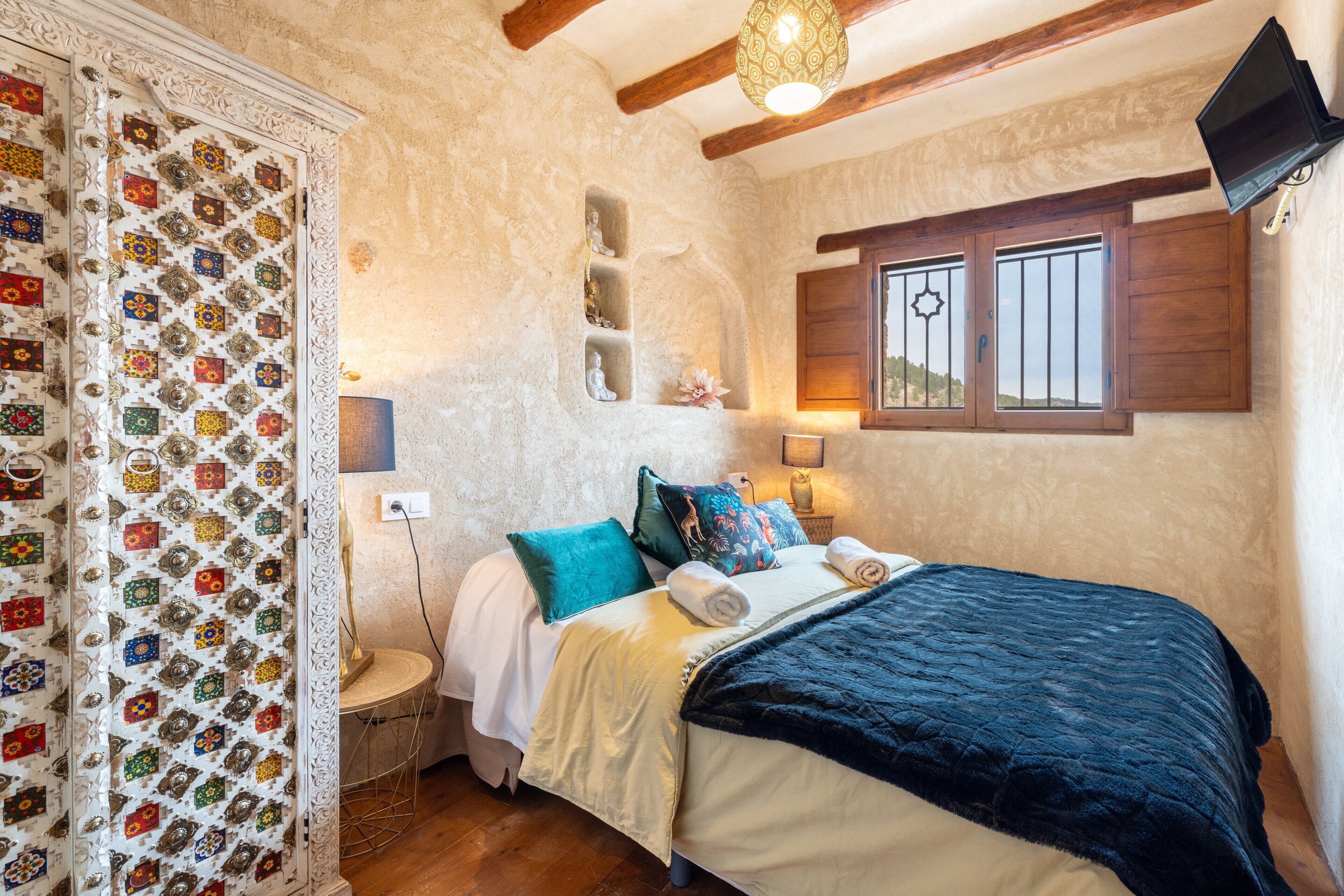 Une chambre à coucher avec un lit, des oreillers, une table de chevet, un téléviseur fixé au mur et une fenêtre avec des volets en bois.