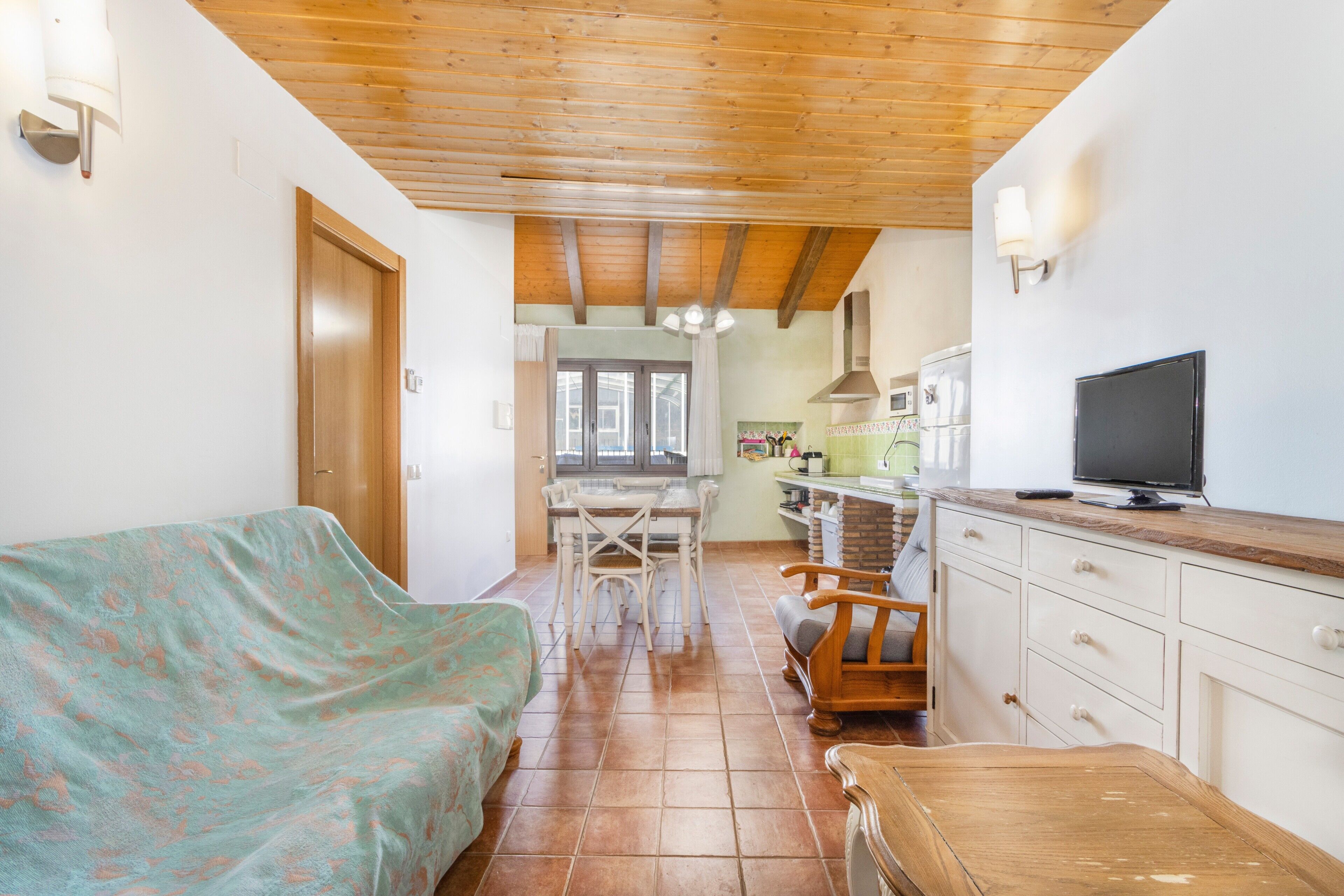 Una habitación con techo de madera, un sofá, una mesa de comedor con sillas, una zona de cocina con televisor y una ventana.