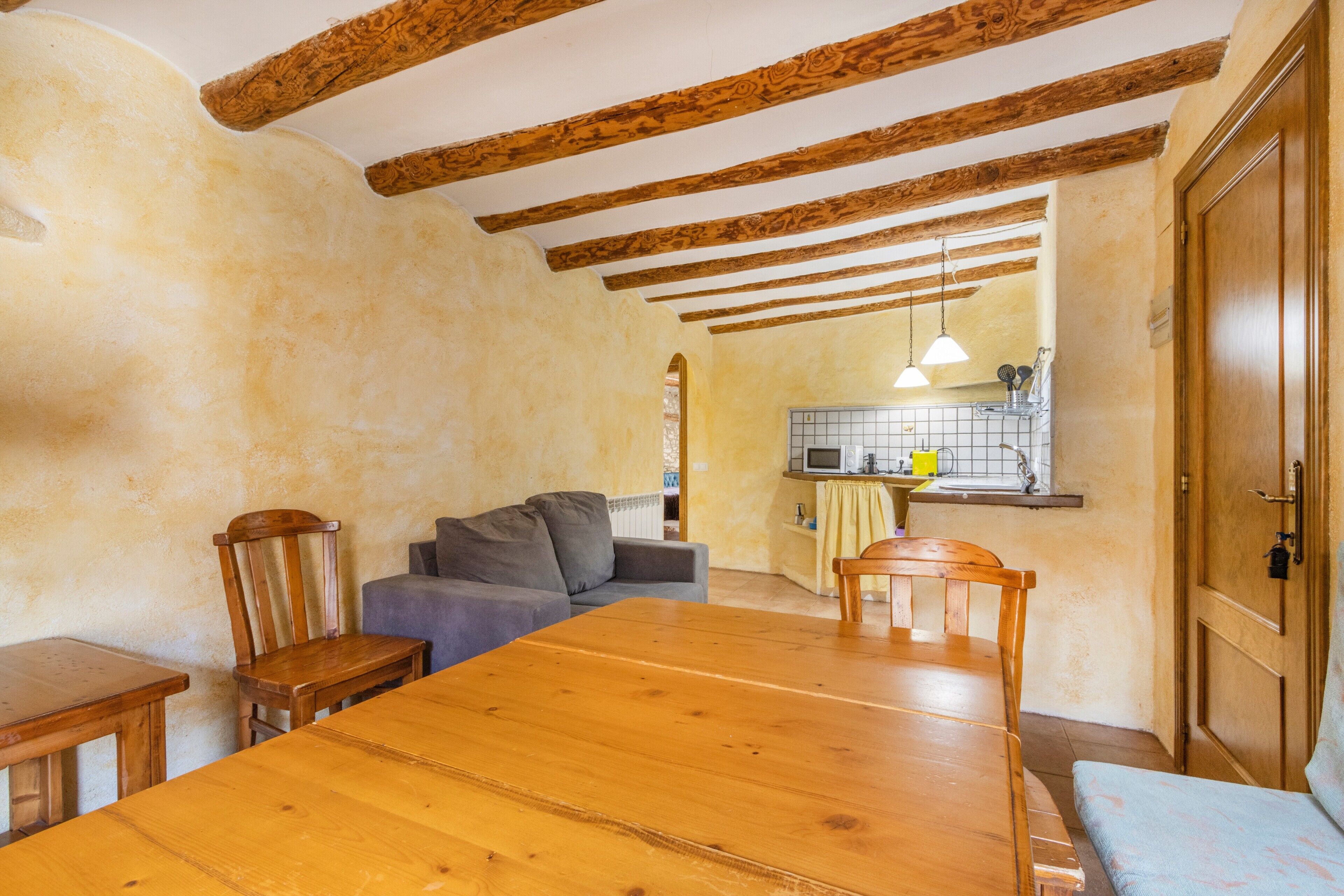 A room with a wooden dining table, two wooden chairs, a sofa, a wooden chair, a wooden bench, a kitchen area with a sink and a window, and a wooden door.