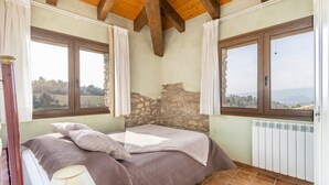Une chambre avec un mur en pierre, un lit recouvert d’une courtepointe marron, deux fenêtres avec des rideaux et une vue sur la campagne.