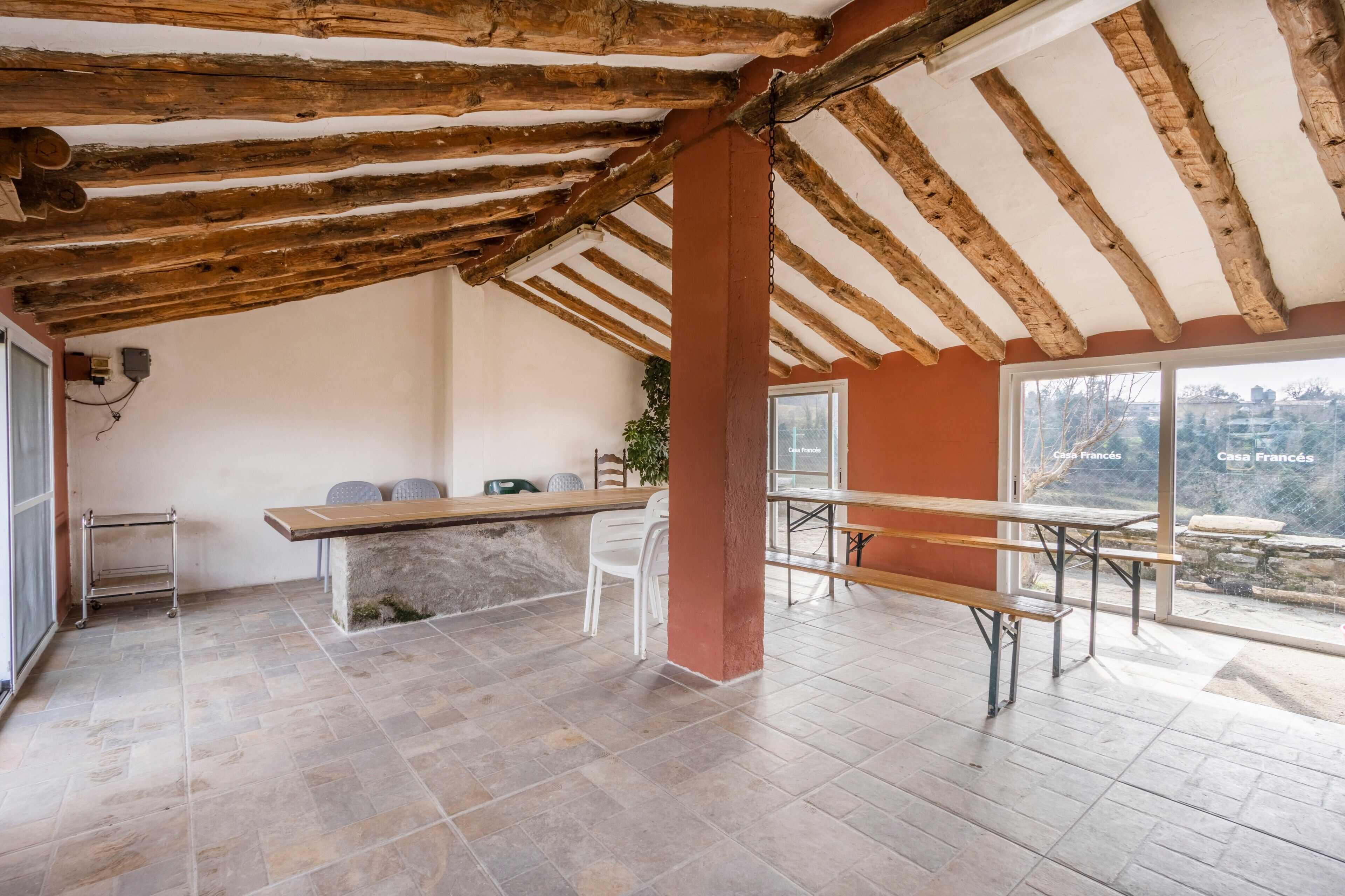 Um quarto espaçoso com teto alto, vigas de madeira aparentes, uma grande janela com vista e um banco simples.