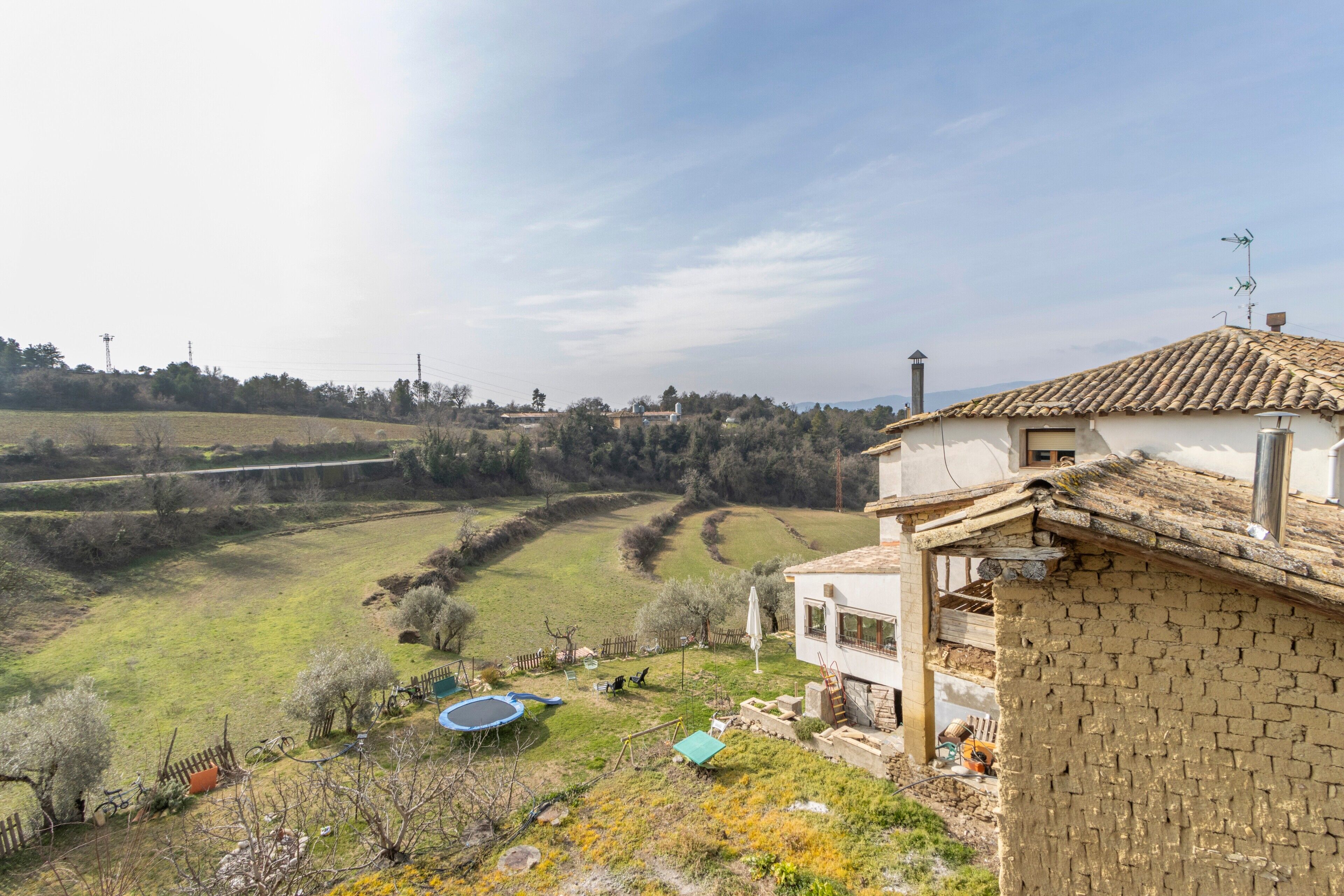 Une maison de campagne avec une façade en briques, un toit en tuiles et une cour clôturée comprenant un trampoline et un petit abri de jardin.