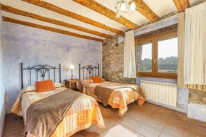 Ein Schlafzimmer mit zwei Betten, einer Steinwand, Holzbalken, einem Fenster mit Vorhängen und Blick ins Grüne.