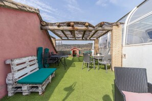 Eine Terrasse mit einer Holzbank, einem Tisch, Stühlen, einer Pergola und einem Gewächshaus.